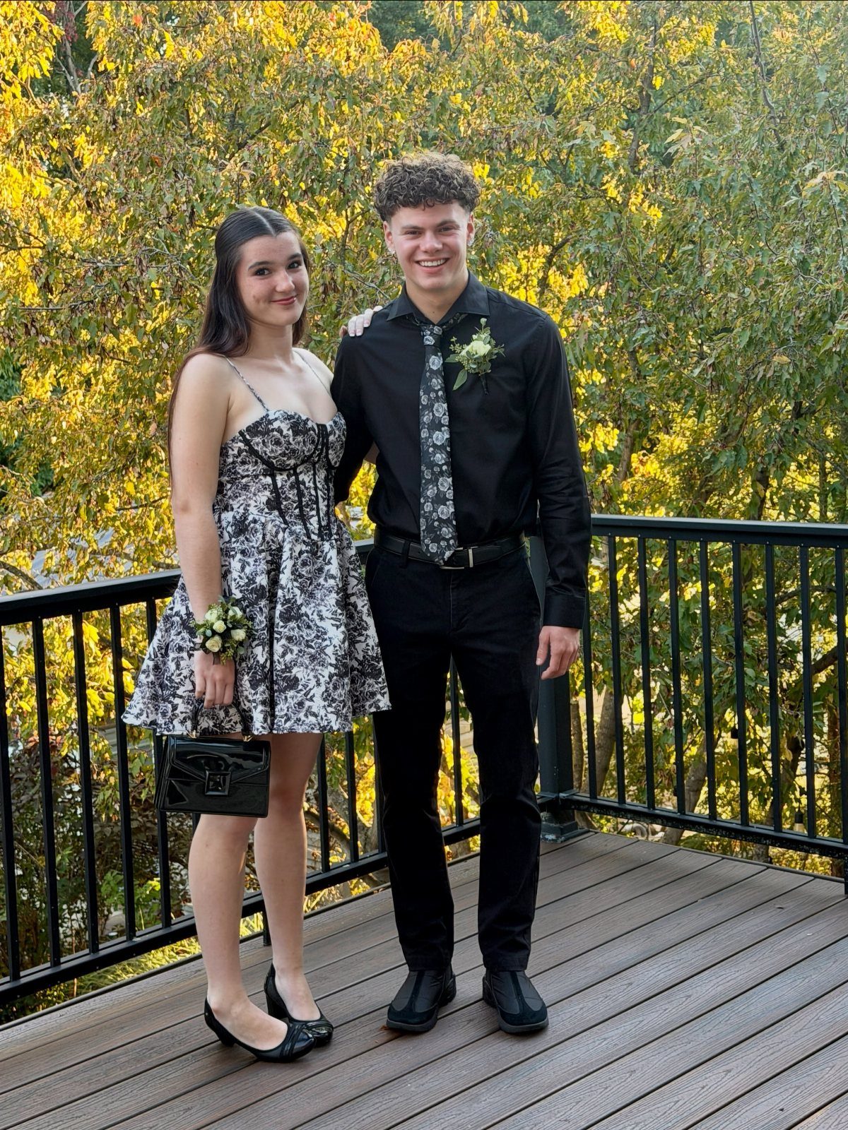 A teenage couple smiling while posing on a wooden deck outdoors with autumn trees.