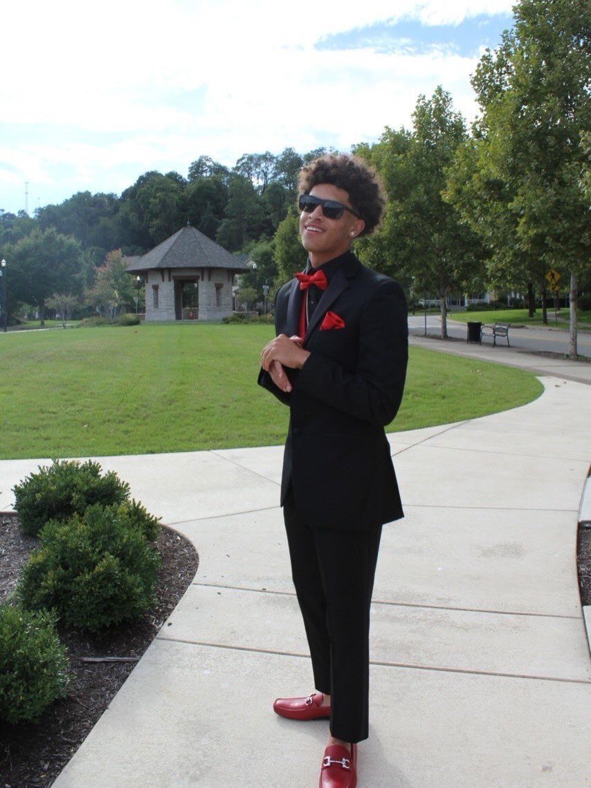 A young man in a black suit and red bowtie wearing red leather loafers on a walkway.