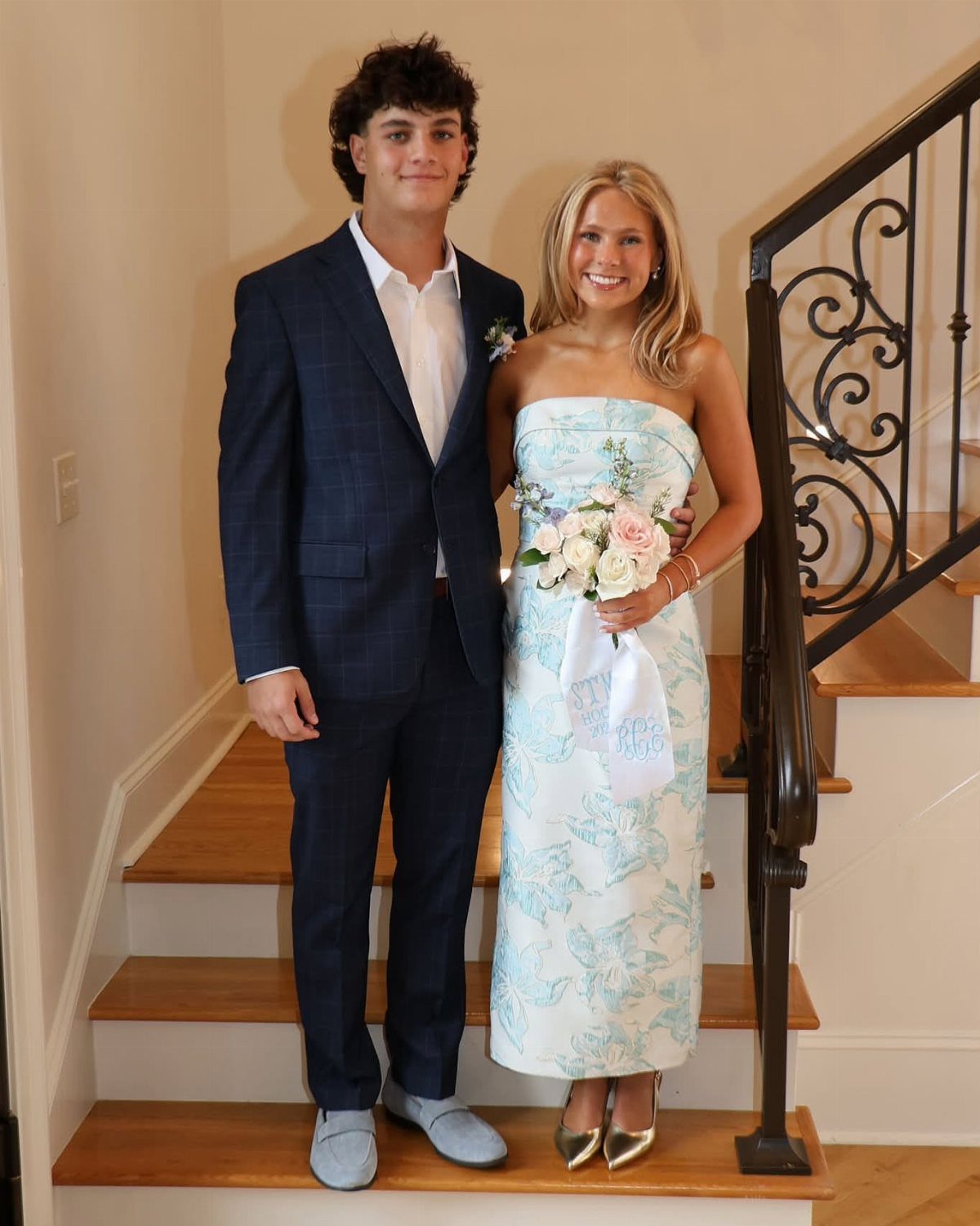 A young man in a navy suit and a woman in a floral dress posing on stairs.
