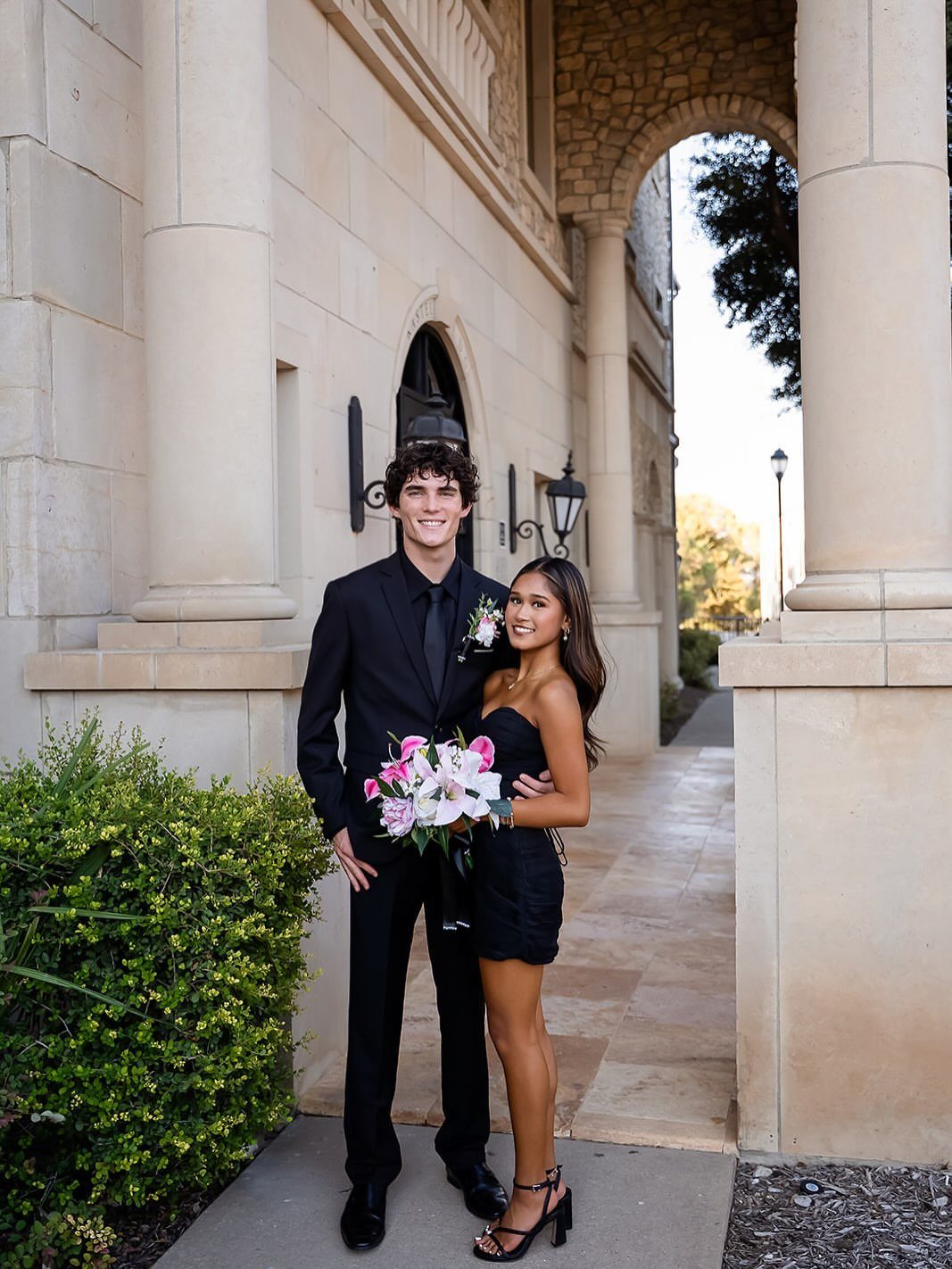 A young man in a black suit and a woman in a black dress posing together.