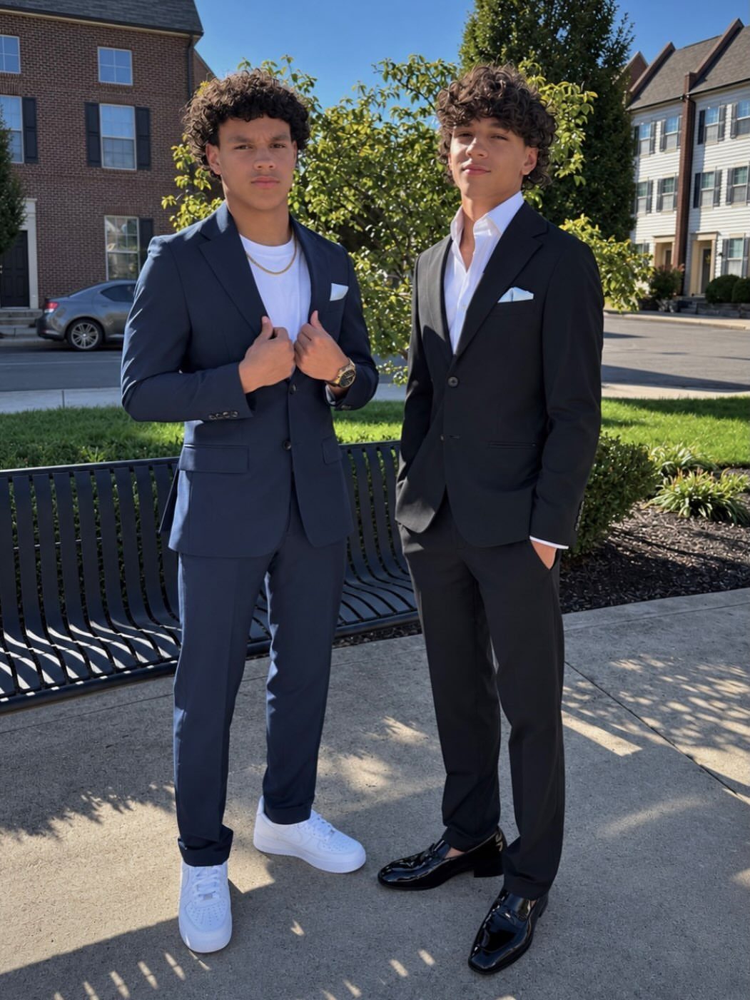Two young men posing in navy and black suits on a sunny sidewalk.