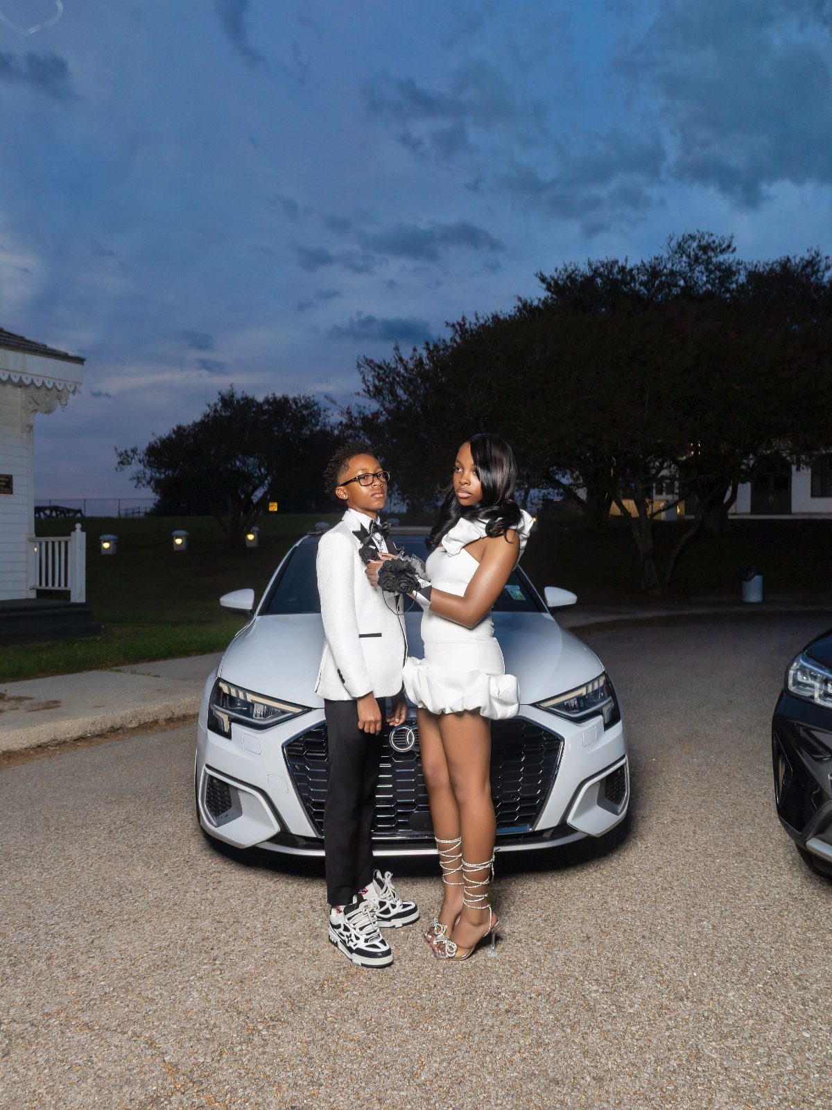 A young man in a white blazer and black trousers posing with a woman by a car.