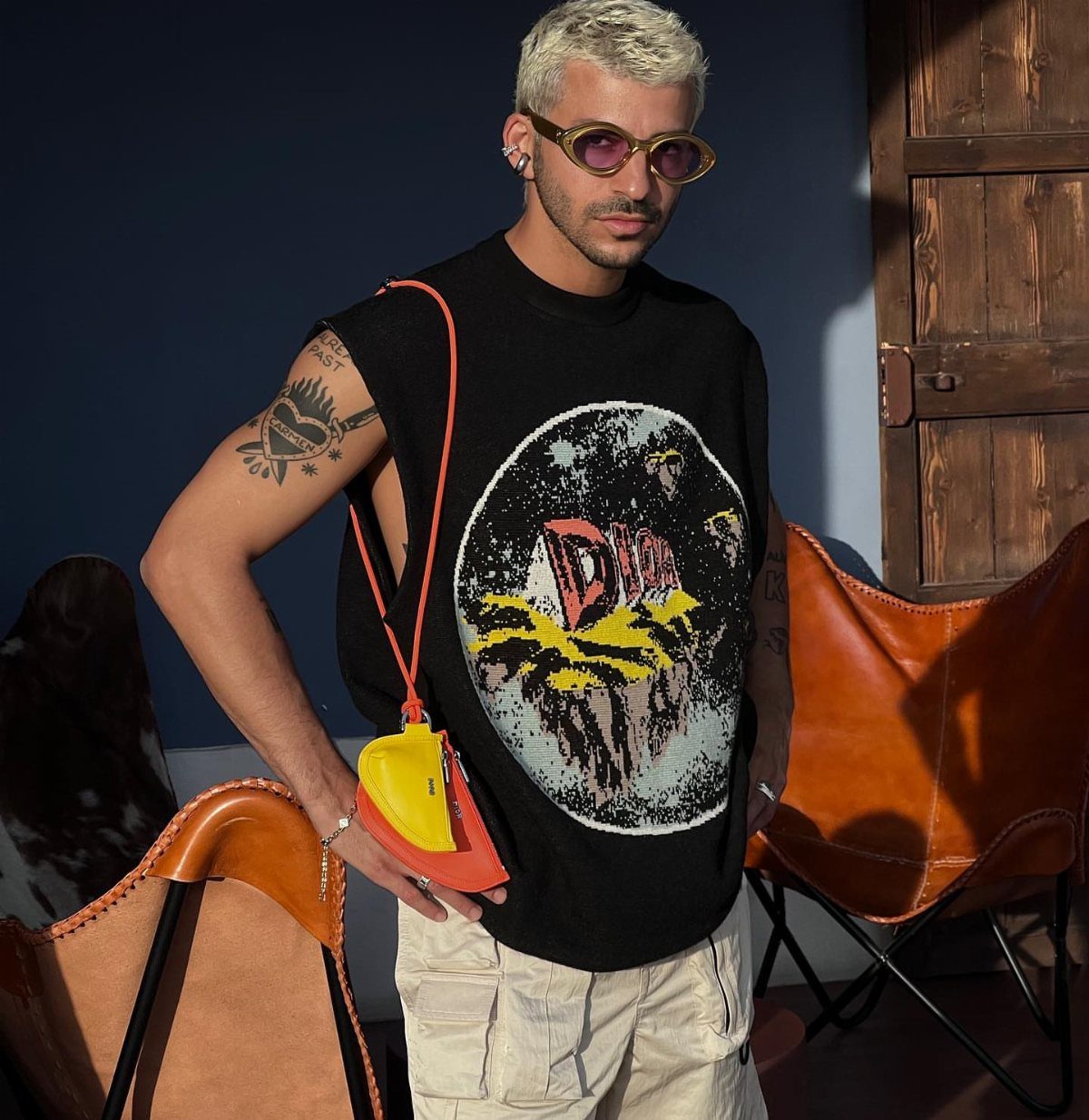 Man with blonde hair wearing yellow sunglasses, a black sleeveless graphic tee, and a small colorful crossbody bag.