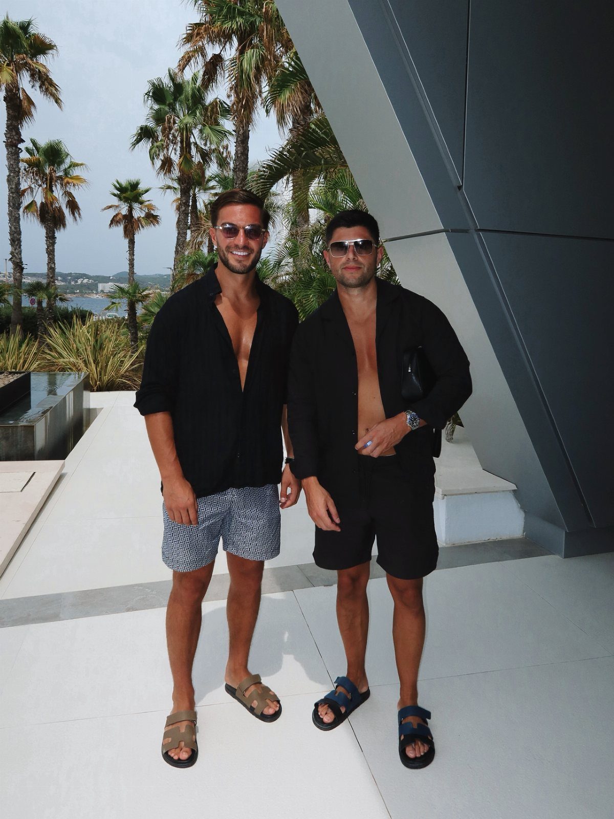 Two men posing in black shirts and summer shorts on a sunny patio.