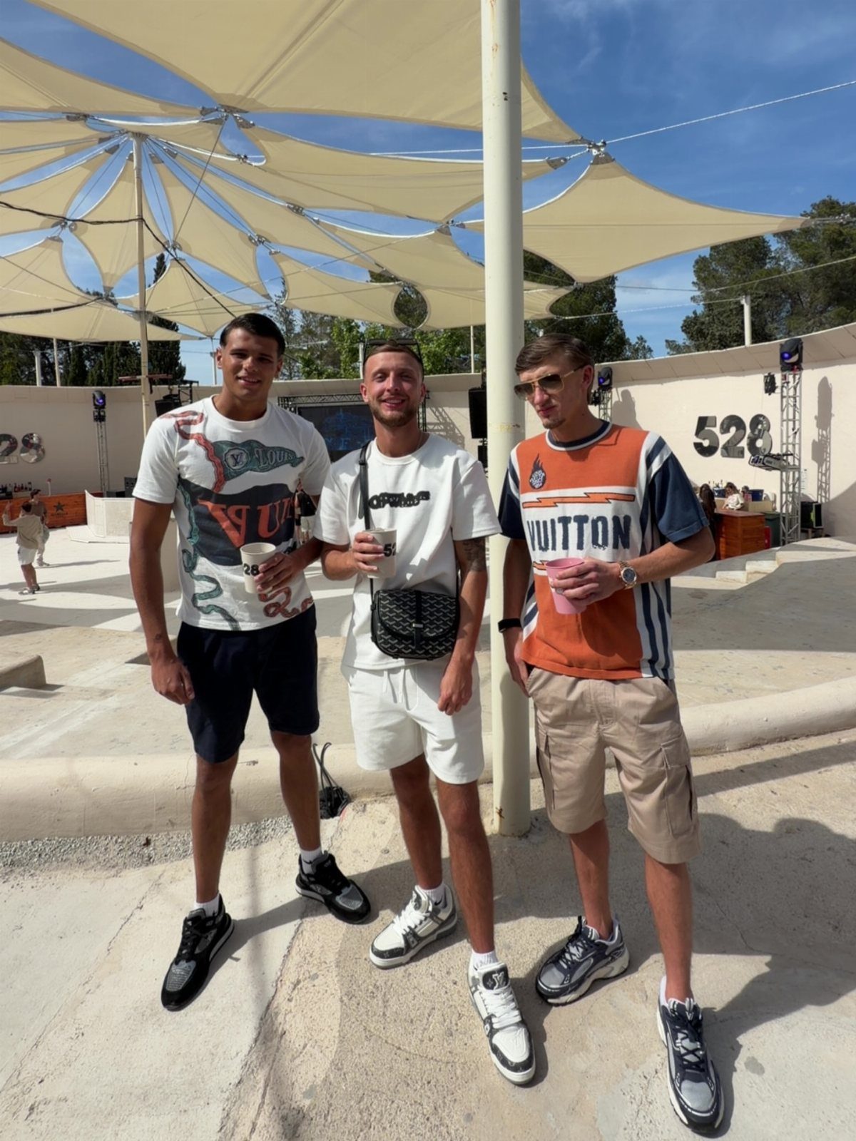 Three men posing in casual summer outfits under a large white canopy outdoors.