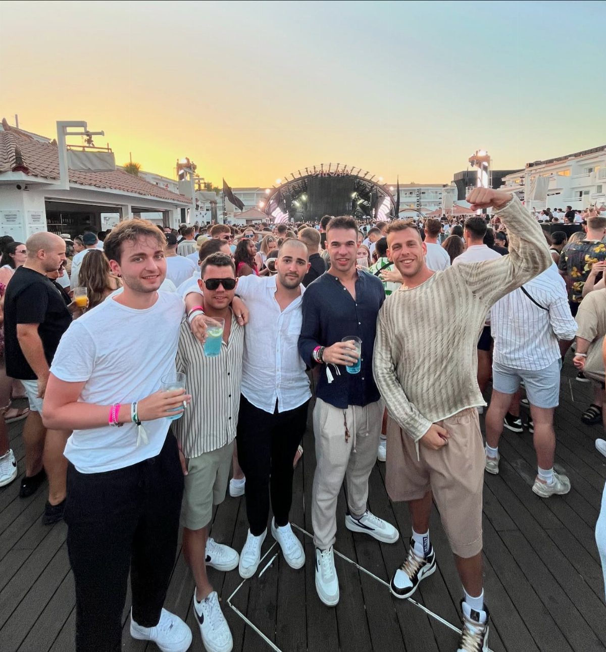 Group of men posing in casual summer outfits at an outdoor beach club during sunset.