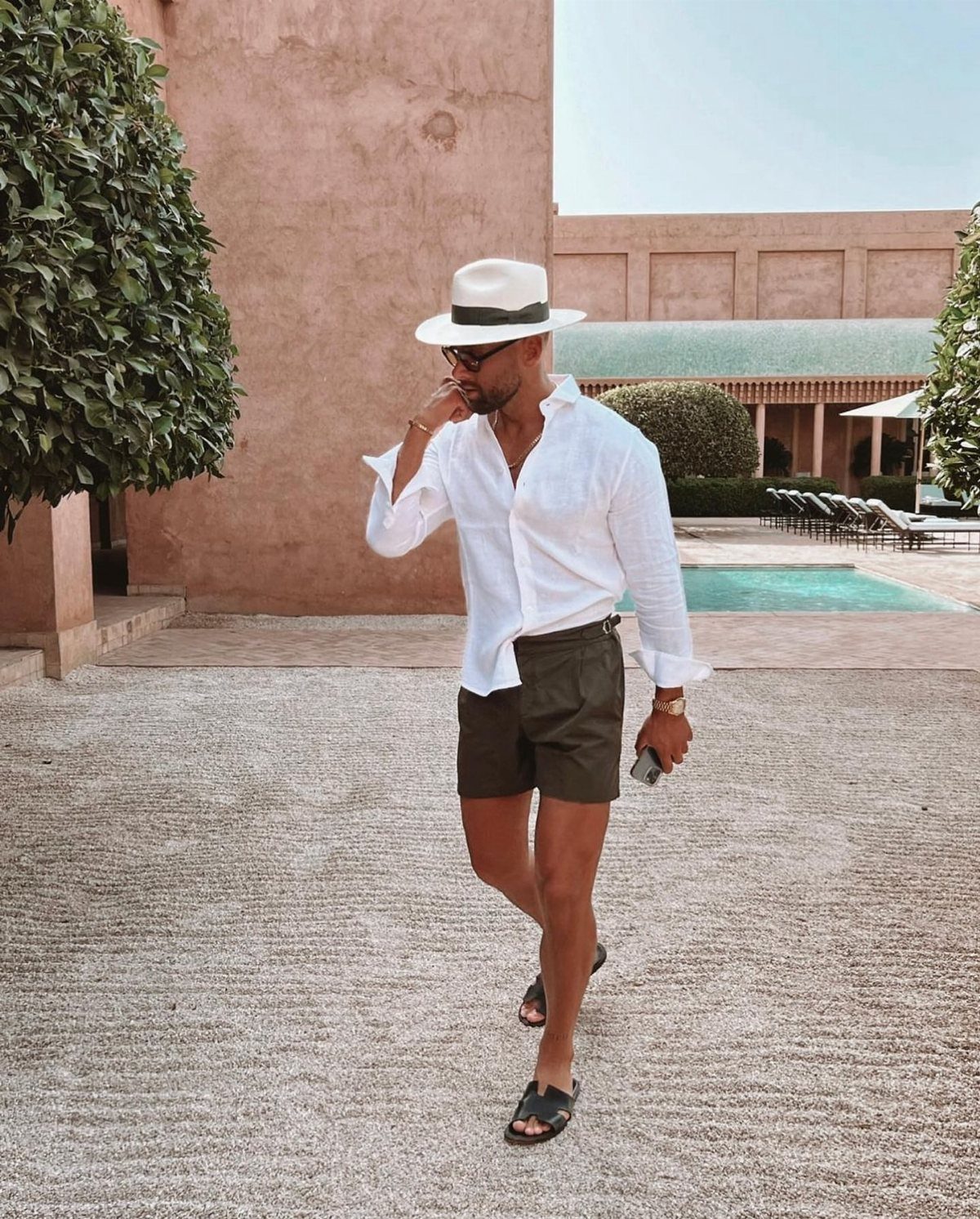 Man in white linen shirt, olive shorts, and fedora walking through a sunny courtyard.