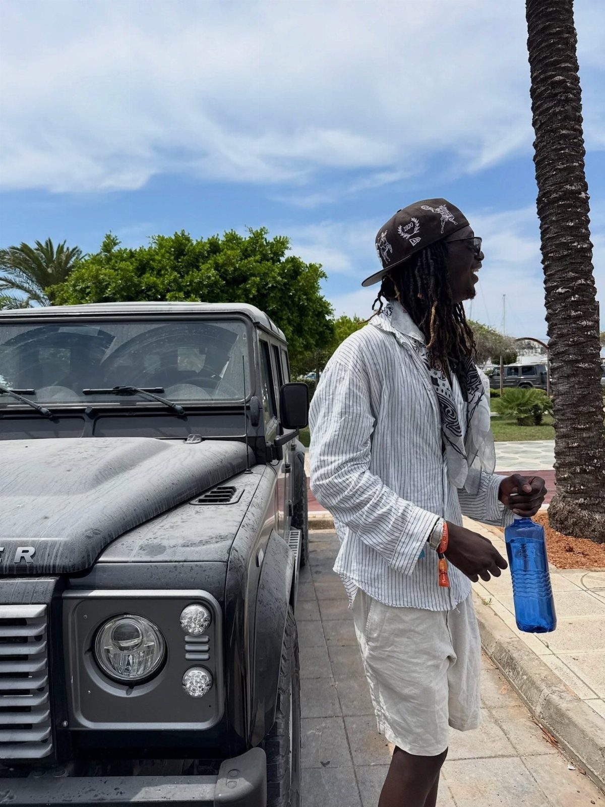 A man in a striped shirt and white shorts stands by a dark SUV under a blue sky.