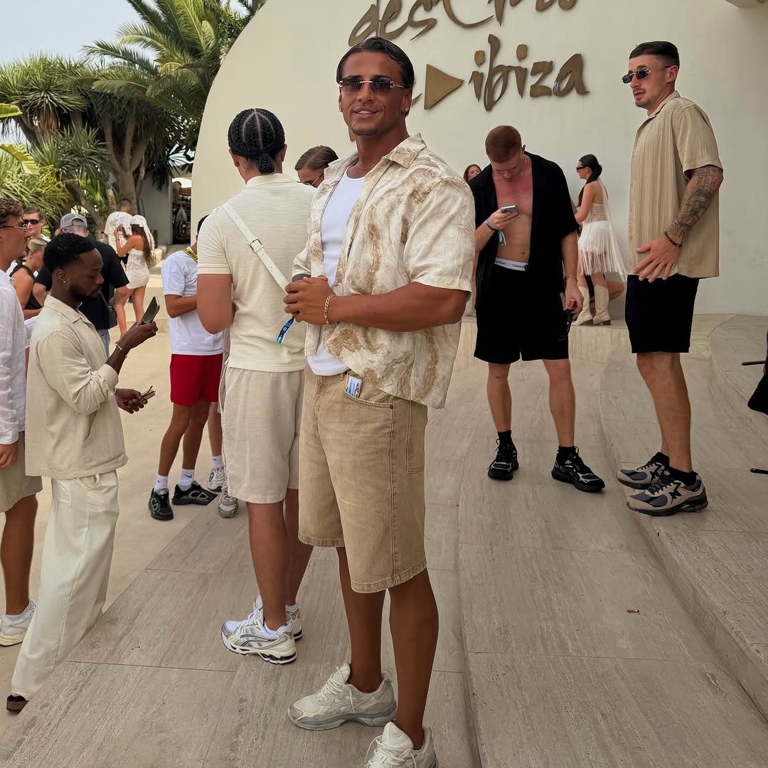 Man in a patterned short-sleeved shirt and tan shorts standing outdoors with other men.