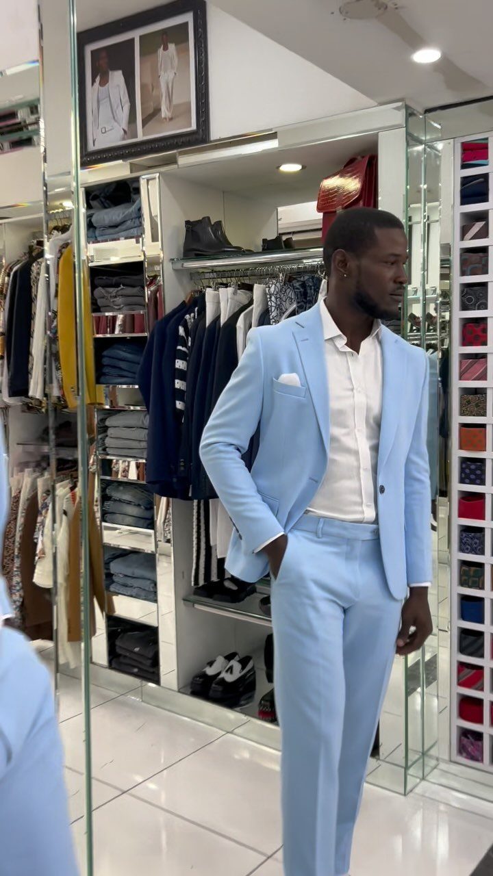 A man posing in a light blue suit and white shirt inside a clothing boutique.