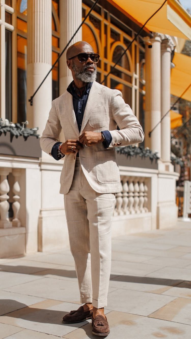 A man in a light beige linen suit and sunglasses standing outdoors near white columns.