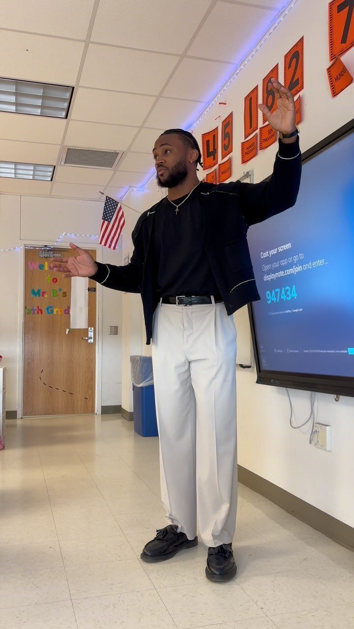Man in a black cardigan and cream trousers gesturing in a classroom setting.
