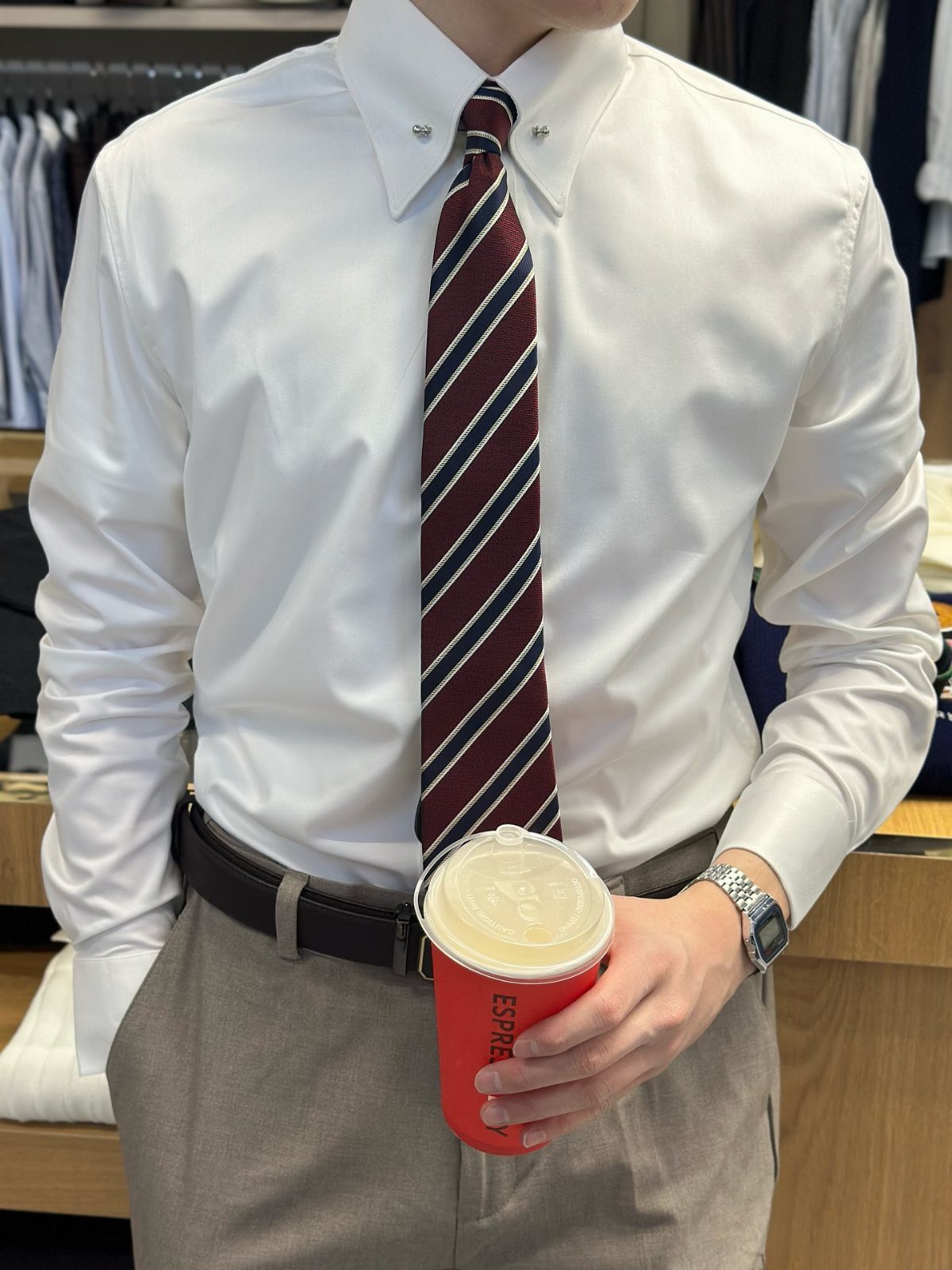 Man wearing a white collared shirt with a striped tie and tan trousers holding coffee.