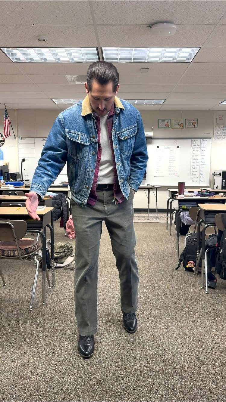 Man wearing a denim jacket, white sweater vest, and maroon shirt in a classroom setting.
