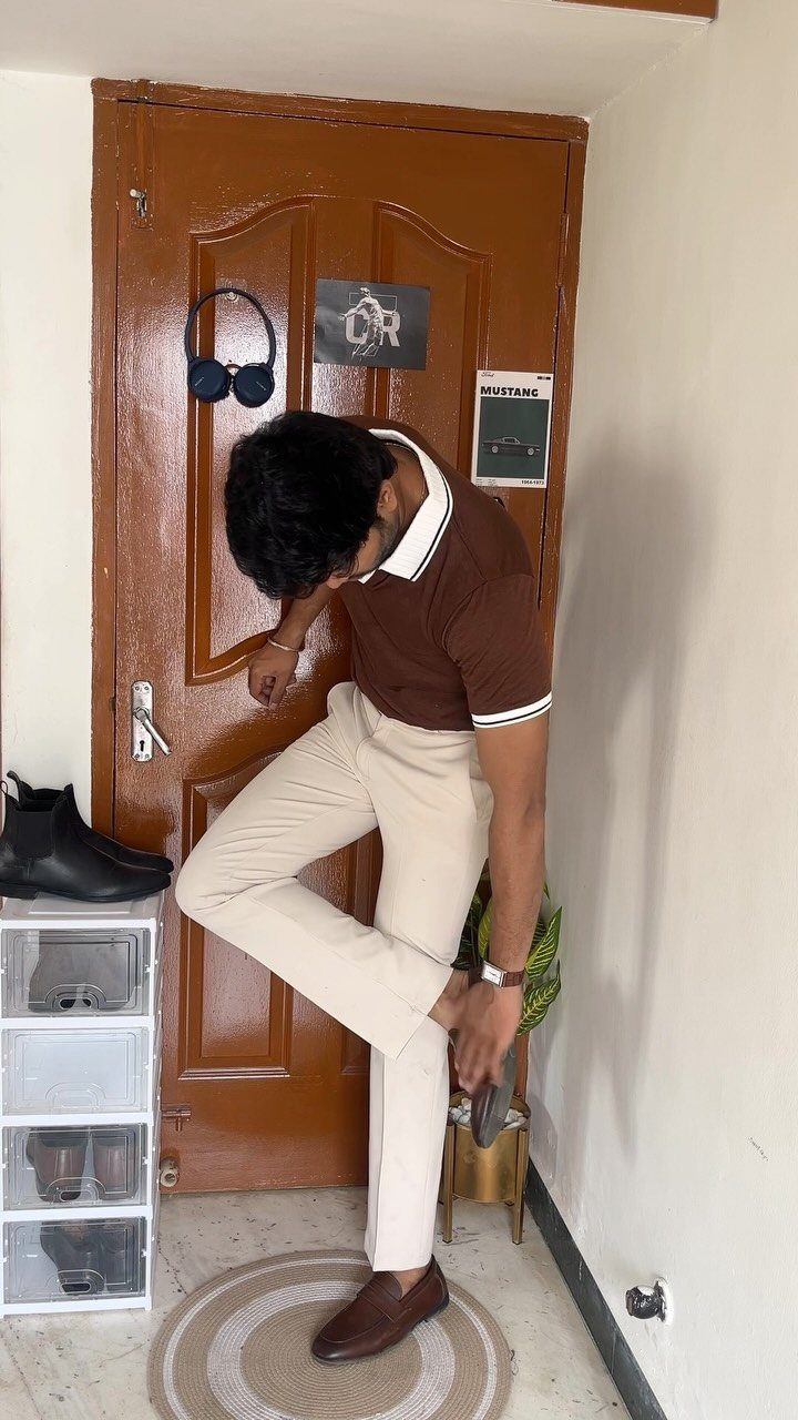 Man in a brown polo and cream trousers putting on brown leather shoes near a door.