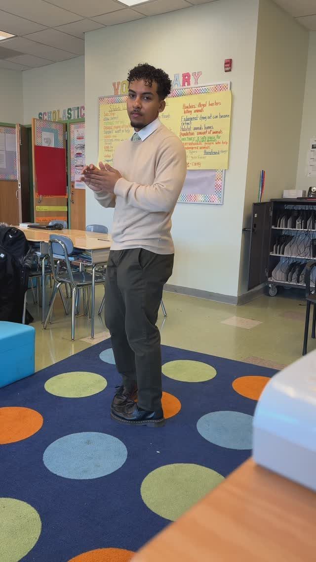 A man wearing a cream sweater over a collared shirt and olive trousers in a classroom.