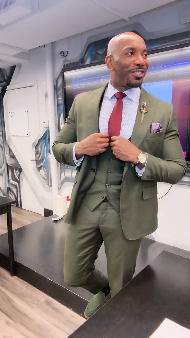 A man smiling while wearing an olive green three-piece suit with a red tie.