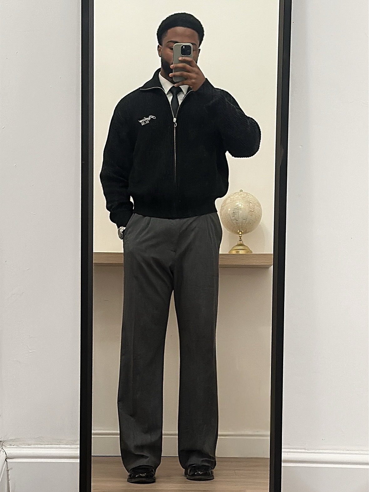 Man in a black jacket, white shirt, tie, and grey trousers posing for a mirror selfie.