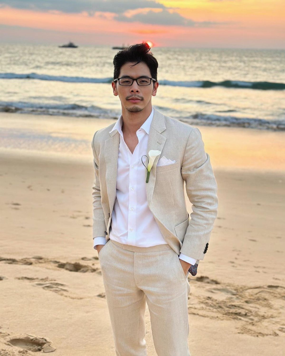 A man in a light-colored linen suit posing on a beach during a sunset.