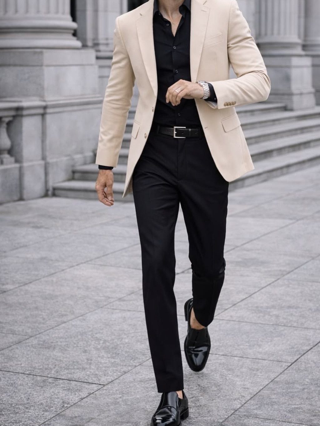 Man walking in a beige blazer, black shirt, black trousers, and black loafers against stone columns.