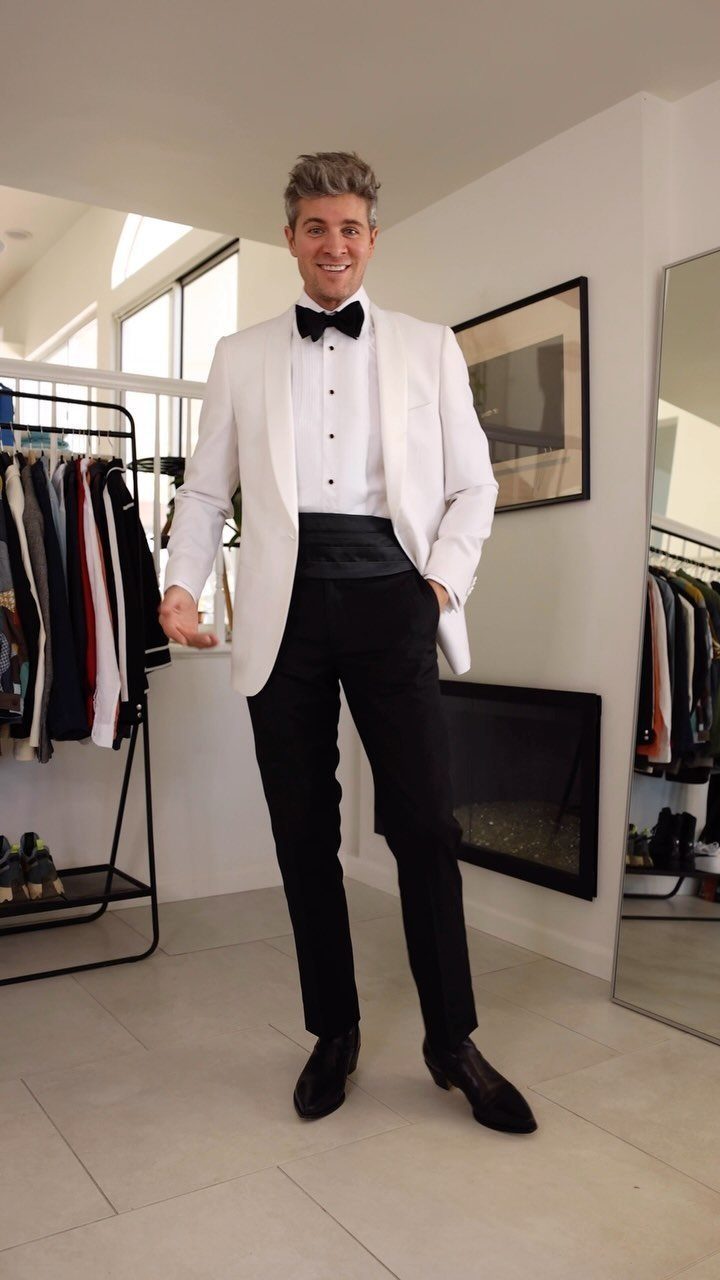 A smiling man wearing a white tuxedo jacket with a black bow tie and black trousers.