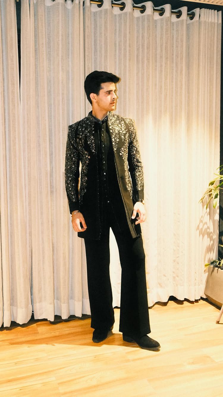 Man wearing a black sequined jacket and wide-leg trousers standing before light curtains.