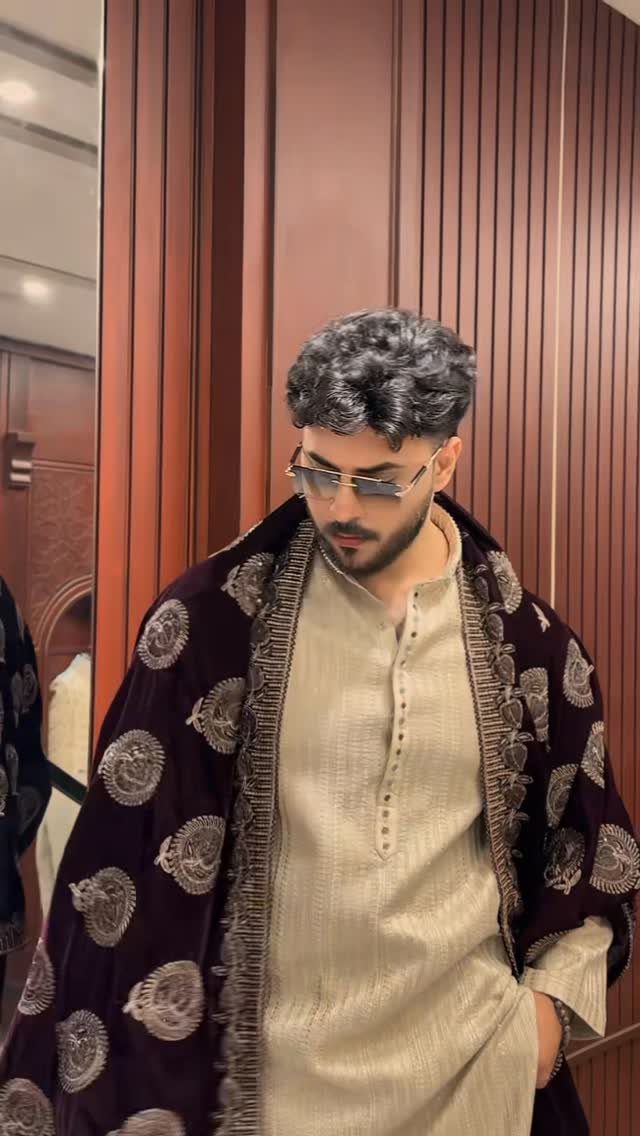 Man wearing a cream tunic and an embroidered maroon velvet shawl against a wooden wall.