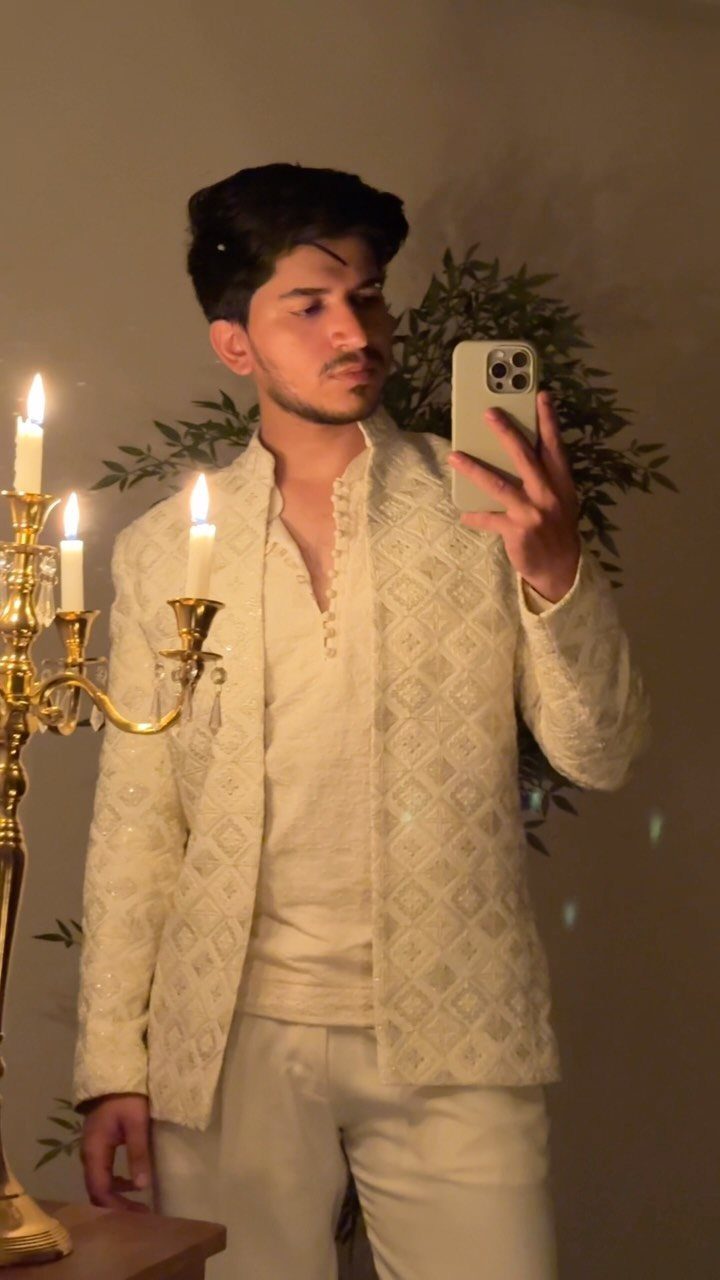 A man posing in a mirror wearing an ivory embroidered traditional Indian engagement outfit.
