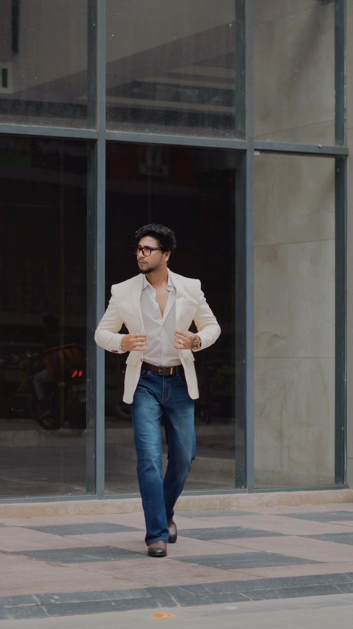A man walking in a white blazer, white shirt, and blue jeans in front of a building.