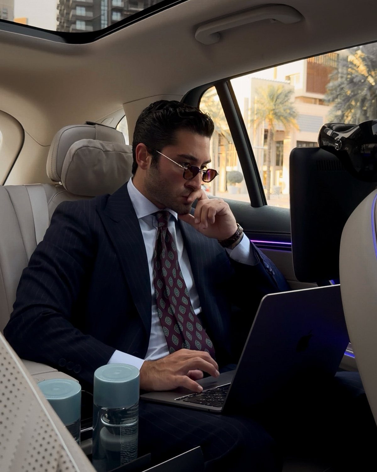 Man in a navy suit and tie working on a laptop inside a luxury car.