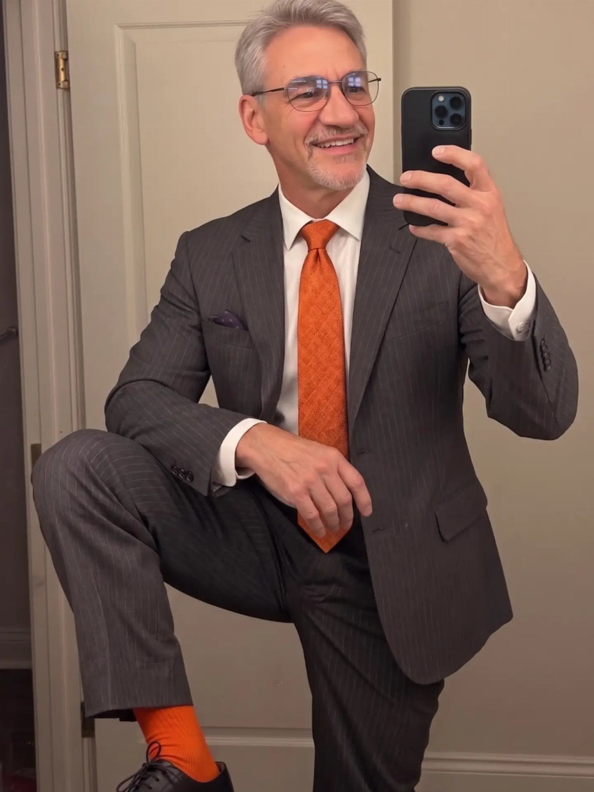 Smiling man in a grey pinstripe suit and orange tie taking a mirror selfie.