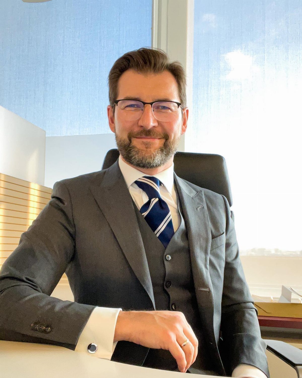 A bearded man in a grey three-piece suit and striped tie sitting at an office desk.