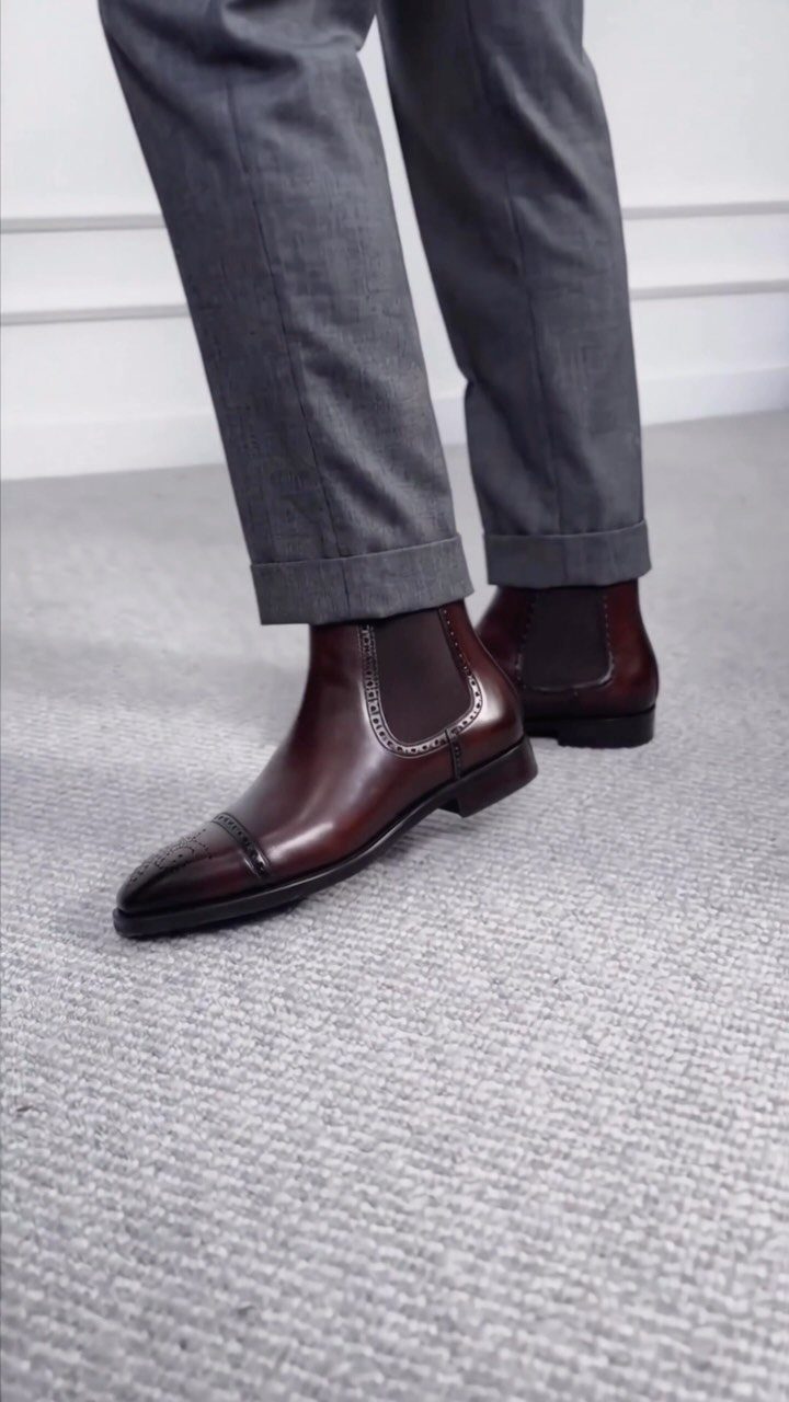 Dark brown leather Chelsea boots worn with grey tailored trousers on a grey carpet.