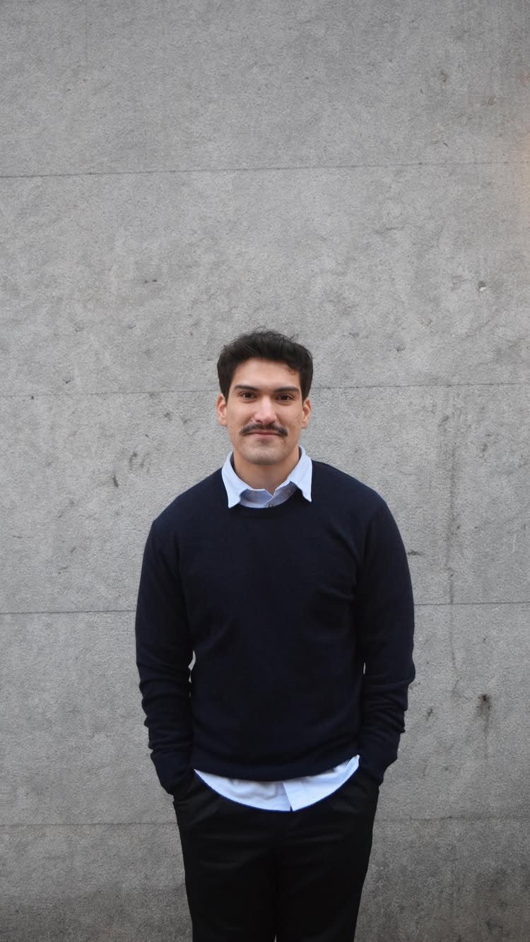 Man wearing a navy sweater over a light blue collared shirt and dark trousers against a grey wall.