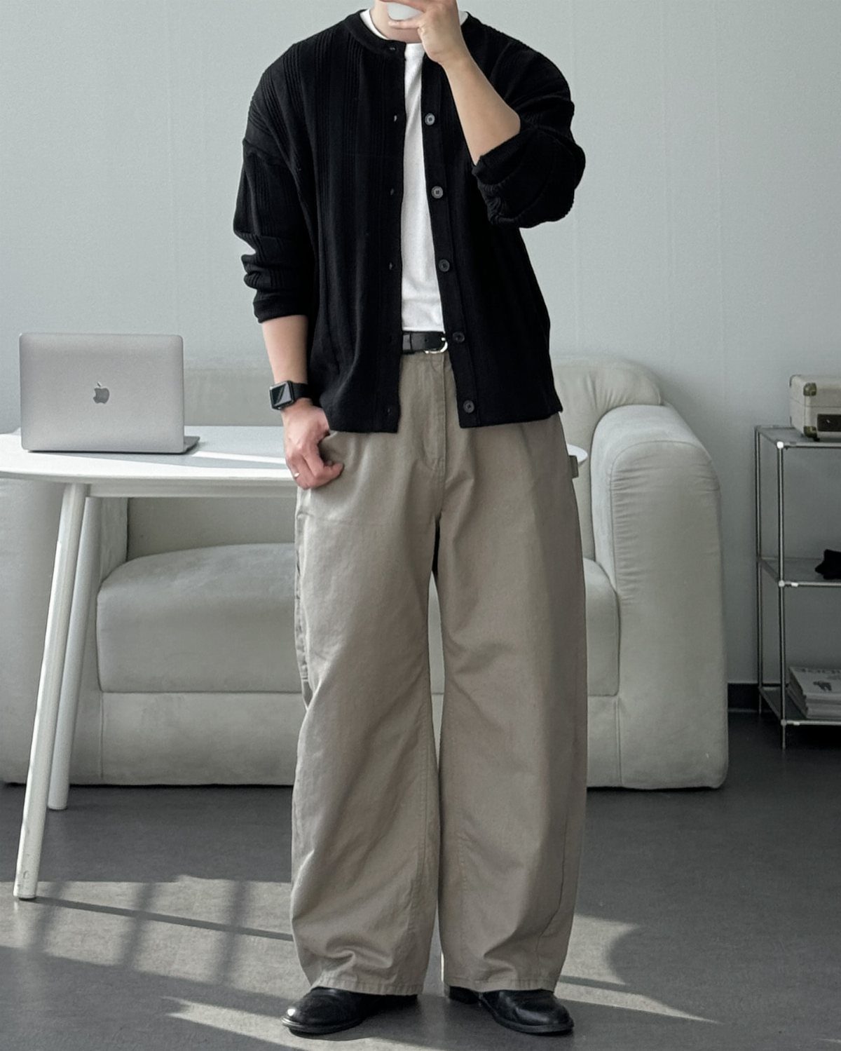 Man wearing a black cardigan, white t-shirt, and wide-leg beige trousers standing in a bright room.
