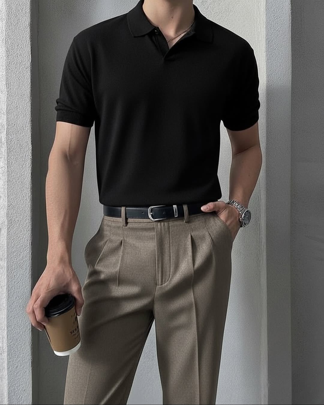 Man in a black polo shirt and khaki pleated trousers holding a coffee cup.