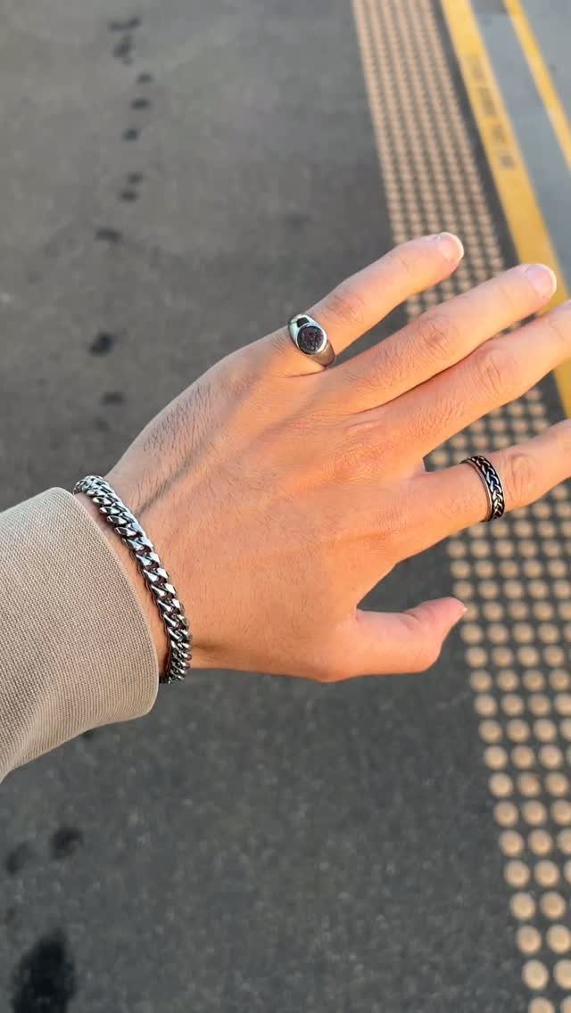A person's hand wearing a chunky silver chain bracelet and two silver rings against asphalt.