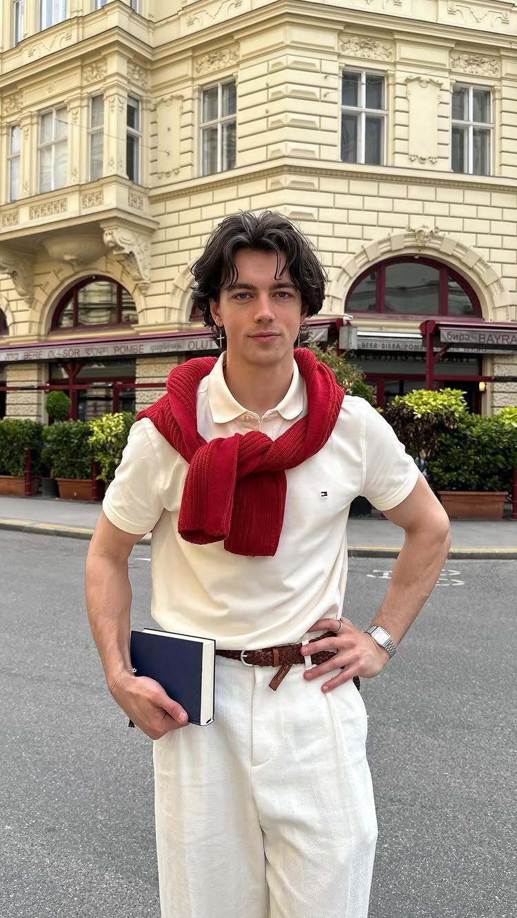A young man in a cream polo, white trousers, and a red sweater draped over his shoulders stands outdoors.