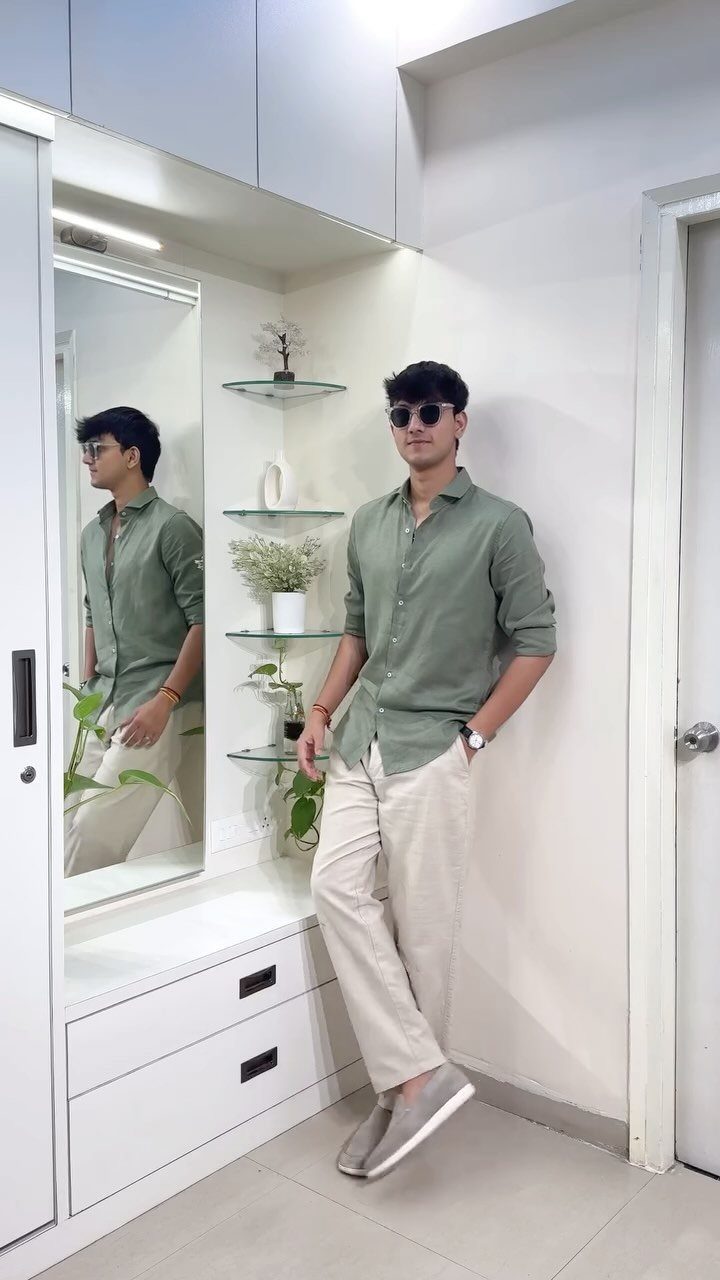 A young man in a green shirt and cream trousers posing against a white wall.