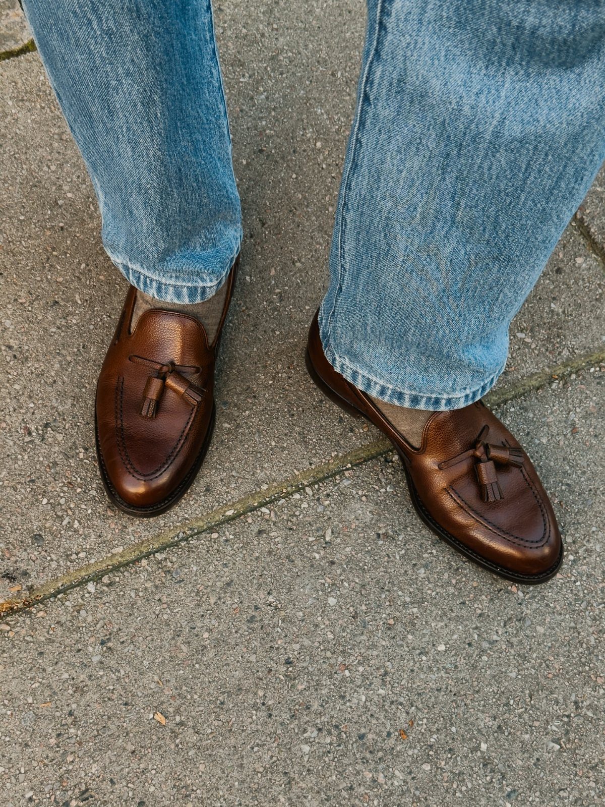 Brown leather tassel loafers paired with light blue denim jeans on a concrete ground.