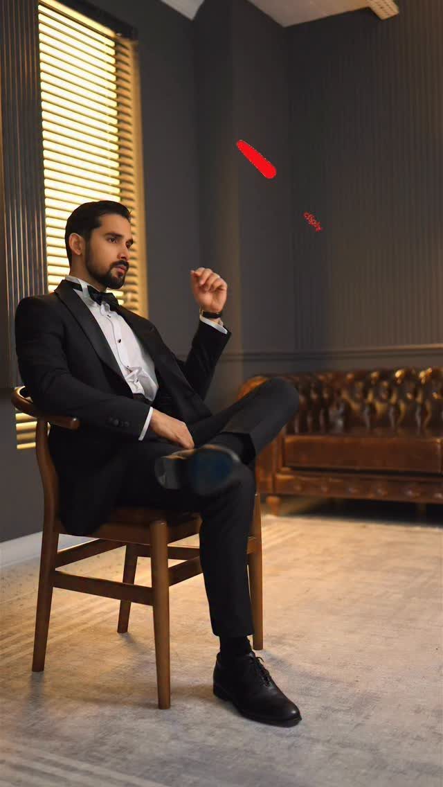 Man in a black tuxedo and bowtie sitting on a wooden chair in a dark room.
