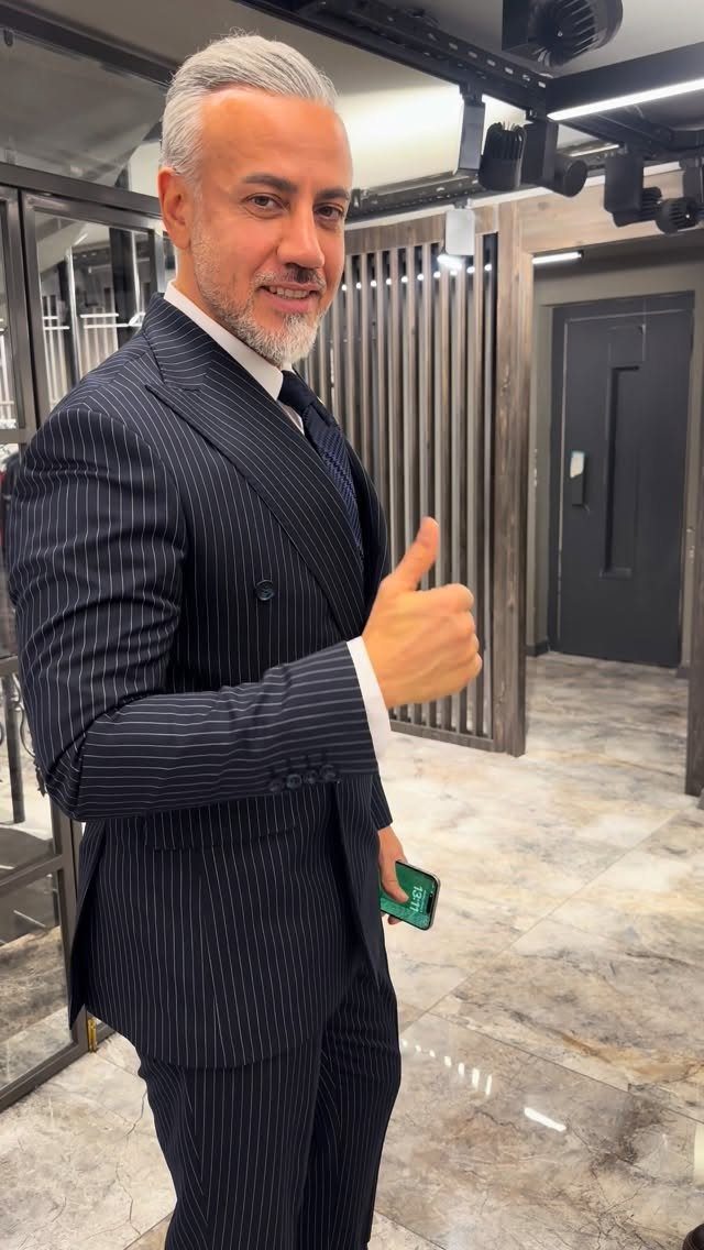 A smiling man with grey hair wearing a navy pinstripe double-breasted suit and giving a thumbs up.