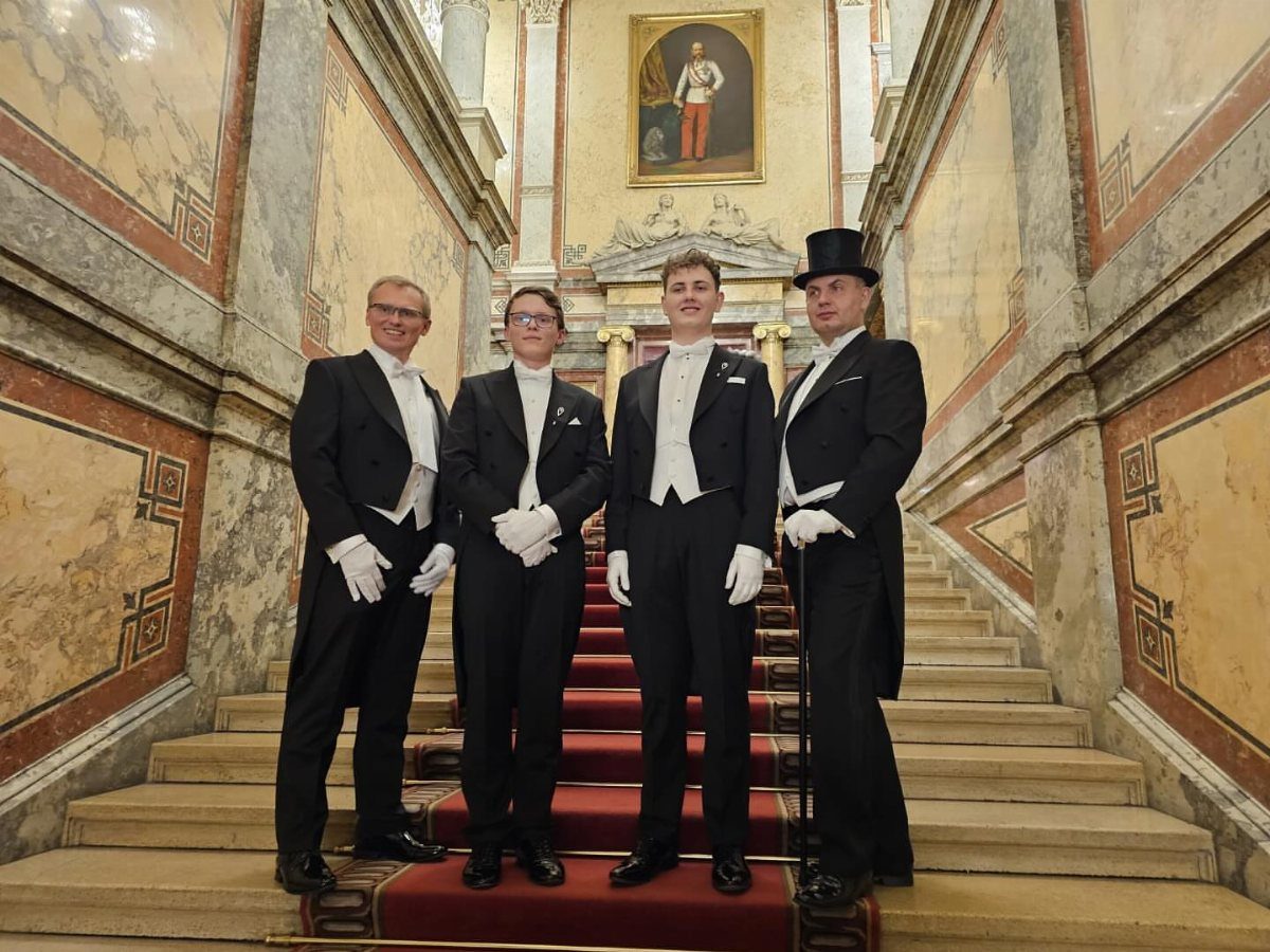 Four people posing on a grand staircase wearing formal white tie attire and white gloves.