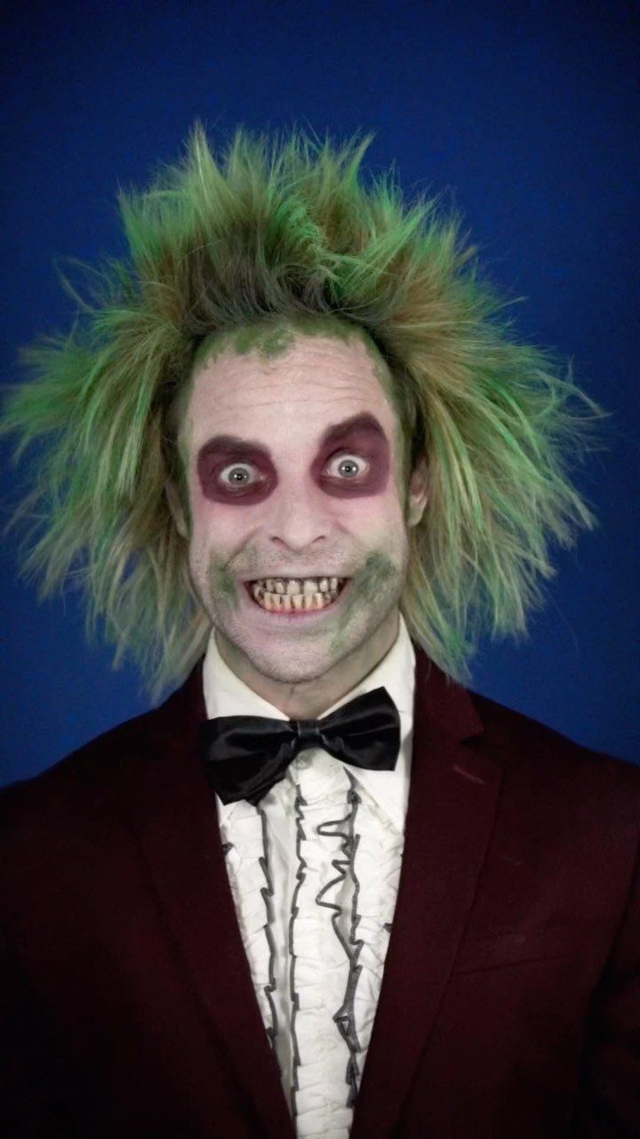 Man in Beetlejuice costume with wild green hair and dark eye makeup smiling against a blue background.