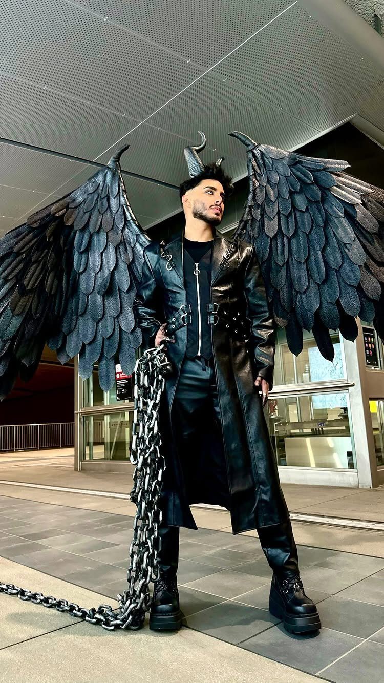 Man in black leather outfit with large dark wings and silver chains standing outdoors.