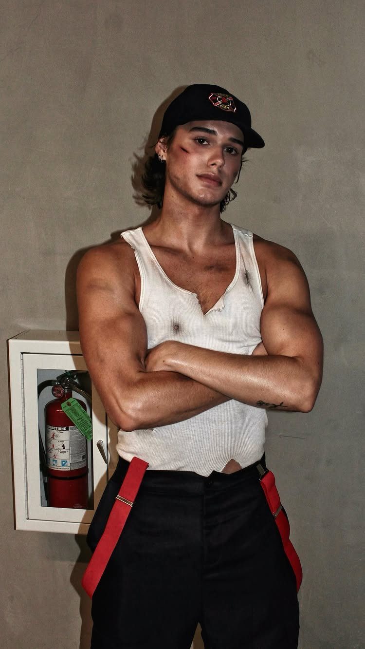 Man in a firefighter costume wearing a white tank top and red suspenders.