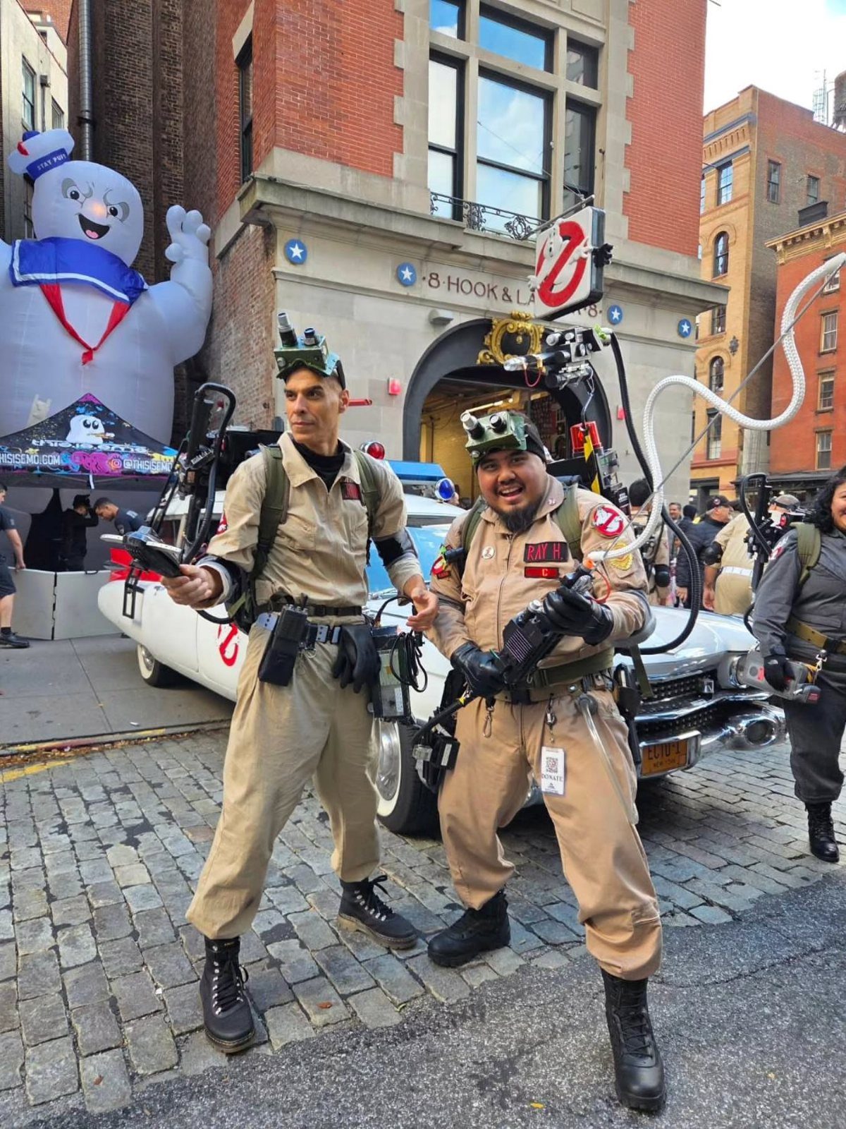 Two men in Ghostbusters jumpsuits posing with an Ecto-1 car and inflatable marshmallow man.