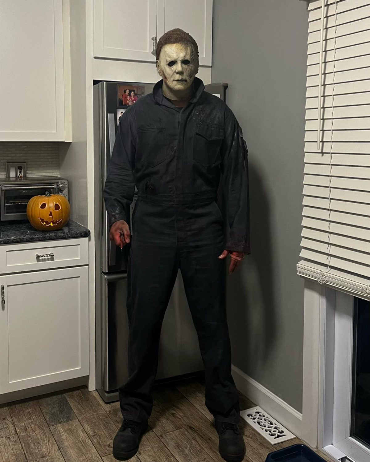 Man in a Michael Myers mask and dark coveralls standing in a kitchen.