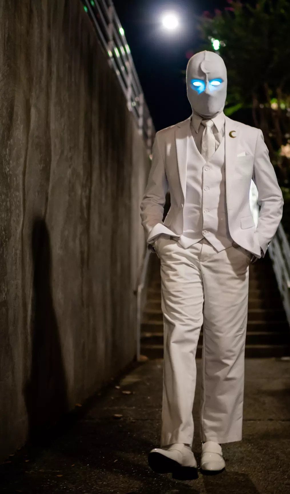 Person in an all-white suit and glowing blue-eyed mask standing by a concrete wall at night.