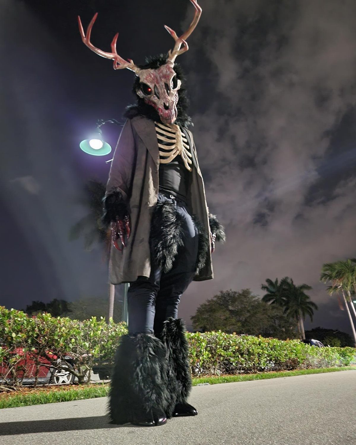 Person in a dark, furry creature costume with antlers and a skull mask standing outdoors at night.